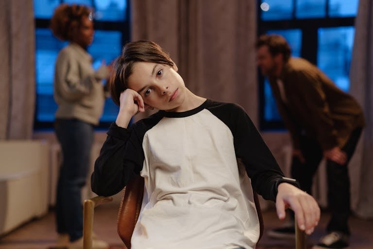 A young woman with short hair and a pensive expression sitting in a dimly lit room with blurred background figures.